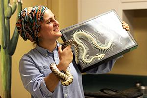 Educator Michelle has a snake wrapped around her left arm and is holding a display case containing a snake skeleton in her right hand